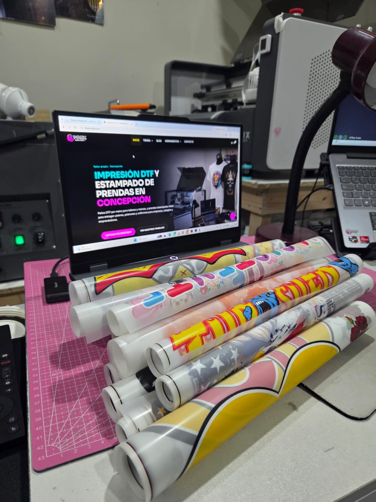 Paños de DTF impresos y enrollados sobre la mesa de trabajo del taller, junto a un notebook que muestra el sitio de Fénix Creations.